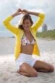 Pretty woman in bikini top, white pants and yellow jacket, kneeling on sand, smiling