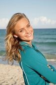 Pretty blonde woman in green zipper jacket standing on beach with arms crossed, smiling