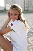 Portrait of pretty blonde woman wearing zipper jacket sitting on beach, smiling