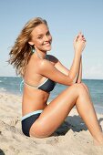 Portrait of beautiful blonde woman wearing gray bikini sitting on beach, smiling