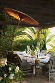 Sommerlich gedeckter Tisch mit Rattanstühlen und asiatischem Sonnenschirm auf Terrasse, im Hintergrund Palmen