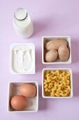 Bottle of milk, egg, potatoes, pasta and butter in serving dish against pink background