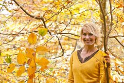 Cheerful blonde woman wearing yellow sweater smiling and holding a branch in autumn forest