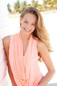 Portrait of happy blonde woman in apricot coloured cowl neck top leaning on wall, smiling