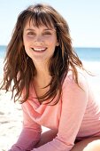 Portrait of pretty woman with brown hair wearing pink shirt standing on beach, smiling