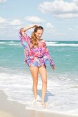 Pretty blonde woman wearing colourful transparent blouse walking on beach, smiling