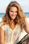 blonde Frau im Netzpulli am Strand, lächelt im Kamera