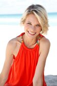 Portrait of pretty blonde woman wearing an orange top standing on beach, smiling