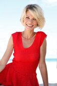 Portrait of pretty blonde woman with short hair wearing red top sitting on beach, smiling