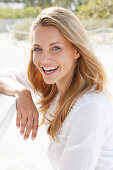 Portrait of beautiful blonde woman wearing white tunic blouse standing on beach, smiling