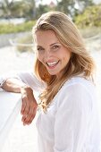 Portrait of beautiful blonde woman wearing white tunic blouse standing on beach, smiling