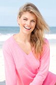 Portrait of pretty blonde woman wearing pink sweater sitting on beach, smiling