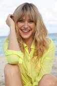 Portrait of cheerful blonde woman wearing yellow shirt sitting on beach, smiling