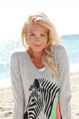blonde Frau im grauen Pulli lächelt in Kamera, am Strand