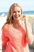 Blonde Frau im orangefarbenen Pulli am Strand, lächelt in die Kamera