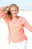 Blonde Frau im orangefarbenen Pulli am Strand, lächelt in die Kamera