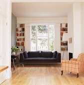 Schwarzes Sofa unter Fenster und antiker Sessel mit kariertem Bezug im offenen Wohnzimmer
