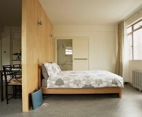 Modern bed with wooden frame in front of a wooden room divider and view into a bath ensuite and chairs behind the divider