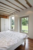 Schlafraum im renovierten Haus mit alten Holzbalken und abgehängter Decke und Terrassentür mit Gartenblick
