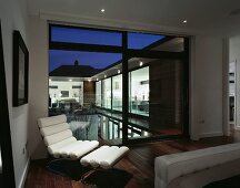 Modern, white leather lounger with foot rest in front of a terrace window and a view of a pool in the evening light