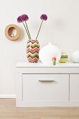 Vases and ornamental flowers on a chest of drawers against a wall decorated with spotted wallpaper