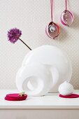 An ornamental flower in a white vase on a chest of drawers against a wall decorated with spotted wallpaper