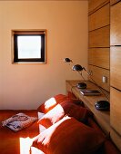 Orange cushions on a bed under a small window and bedside lamps on a wooden shelf