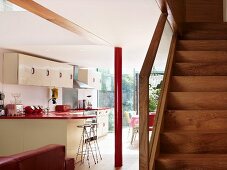 Schnörkellose Treppe aus Holz neben offener Küche mit weisser Schrankfront und rot lackierter Metallstütze