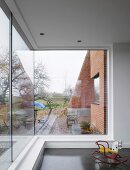 Small, colourful rocking horse in front of large corner window with view of a garden and brick walls