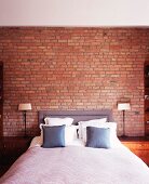 Double bed with upholstered head and scatter cushions flanked by graceful bedside lamps in front of brick wall