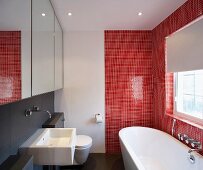 Modern bathroom with bathtub against red mosaic tiles and basin under mirrored cupboard