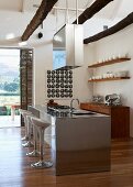Open kitchen with designer barstools at stainless steel kitchen island, suspended extractor hood and white crockery on open wooden shelves