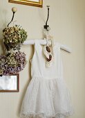 Weisses Babykleid mit Schühchen an einem Wandhaken neben getrockneten Blumensträussen