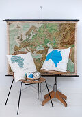 Kissen mit Landkartenmotiv auf Stuhl und Drehstuhl aus Holz vor aufgehängter Landkarte an Wand