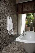 Free-standing bathtub and mosaic tiles on walls and floor in modern bathroom with open doorway leading to courtyard