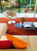 Klassiker Couchtisch und weisser Designer Drehsessel auf rotem flokatiartigem Teppich in zeitgenössischer Architektur mit Glasfassade und Blick in Garten