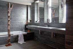 A grey bathroom with stone tiles on the floor and on the walls with a tall wooden sculpture in front of a bathtub
