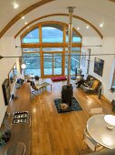 Open-plan living space seen from above with central wood-burning stove below arched ceiling and glass wall with view of wooden terrace