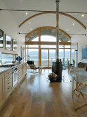 Lange Küchenzeile und zentraler Kaminofen in offenem Wohnraum mit Tonnendach und Blick auf Küstenlandschaft