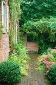 Ländlich gepflasterter Weg im Garten und Rosen vor Wohnhaus mit begrünter Ziegelfassade. Durchgang in weiteren Außenbereich.