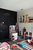 Children's drawings on black wall behind white children's furniture, colourful rug and girls' and boys' toys