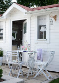 weiße Gartenstühle und Tisch vor Gartenhäuschen mit weiss gestrichener Holzfassade