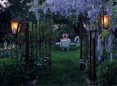 Gartentor unter blau blühender Glyzinienpracht und romantisch gedeckter Gartentisch im Hintergrund