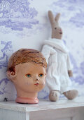 Doll's head and rabbit soft toy on shelf against Toile de Jouy wallpaper