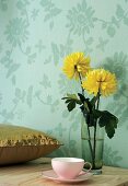 Gelbe Blumen in Trinkglas und Paillettenkissen vor Blumentapete