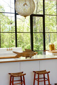 Fish-shaped, wooden ornament on modern kitchen counter and rustic barstools in front of lattice window with view of garden