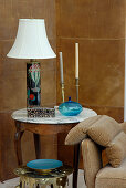 Table lamp with white lampshade and candlesticks on traditional side table in corner of room against wall clad in leather-covered panels