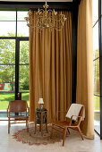 Wooden chairs and side table in front of floor-length, beige curtain in corner of high-ceilinged living room with view of garden