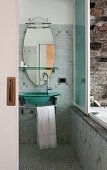 Badezimmer mit Glaswaschbecken und ovalem Spiegel; ein Schiebefenster öffnet den Blick zur aussen platzierten Badewanne