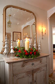 Hall mirror above candle in arrangement of roses and stylised metal fir trees on vintage cabinet
